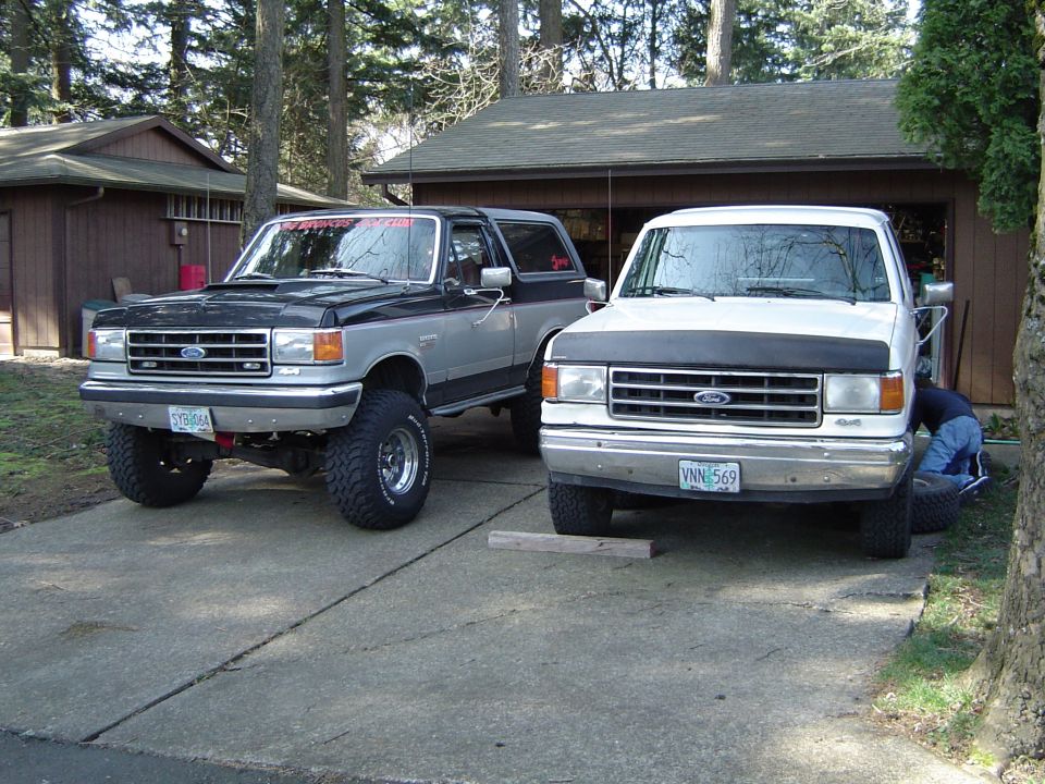 Black/silver lifted Ford Bronco beside white Ford truck on a driveway by a garage.