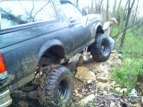1989 Ford Bronco - A dark colored Ford Bronco is shown from the rear passenger side, navigating a rocky, wooded terrain. The vehicle has large, aggressive off…