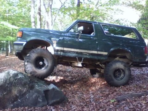 1989 Ford Bronco - A dark green, two-door Ford Bronco is shown from the side, navigating over a large rock on a wooded trail. The vehicle has large, off-road…