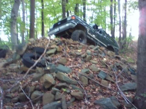 1989 Ford Bronco - A dark green Ford Bronco is tilted on its side, partially submerged in a pile of rocks and debris in a wooded area. The vehicle appears to…
