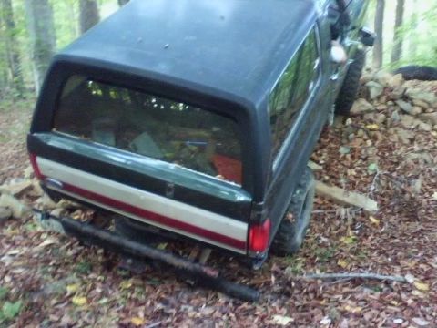 1989 Ford Bronco - The rear passenger side of a dark colored SUV with a camper shell is shown at an angle, resting on a pile of rocks and logs in a wooded are…