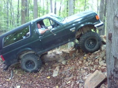 1989 Ford Bronco - A dark green Ford Bronco is navigating a rocky, wooded trail, with its front tires elevated on a pile of rocks. The driver is visible behin…