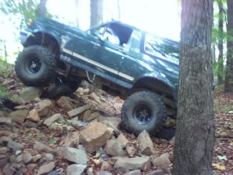 1989 Ford Bronco - A dark green Ford Bronco is navigating a rocky, wooded trail, with its tires angled on large stones. The vehicle is tilted significantly to…