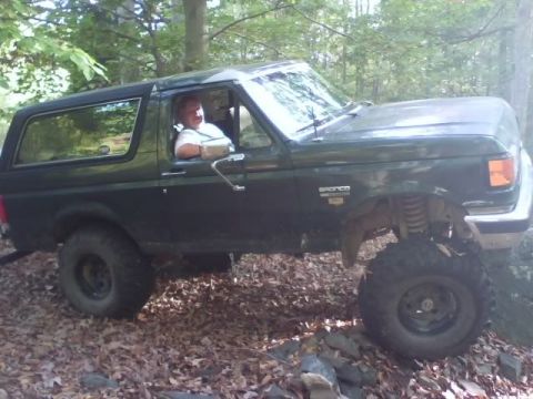 1989 Ford Bronco - A dark green Ford Bronco is shown off-road, with its front passenger side tire elevated on rocks. The driver is visible through the open wi…