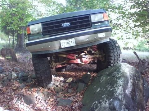 1989 Ford Bronco - A modified dark green Ford Bronco is navigating a rocky, wooded trail, with its front passenger side tire high on a large boulder. The vehi…