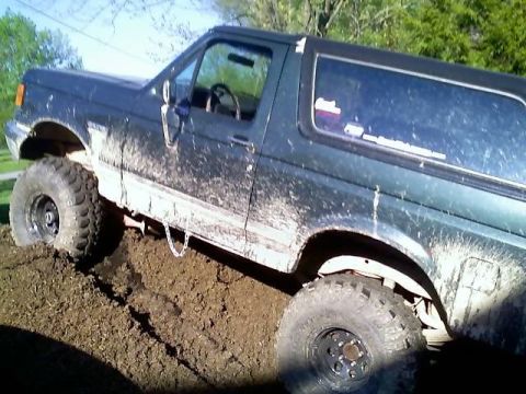 1989 Ford Bronco - A dark green, mud-covered Ford Bronco is stuck in a pile of dirt, with its front tire partially submerged. The vehicle appears to be an SUV…
