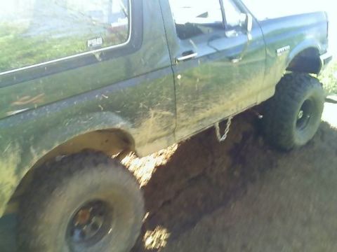 1989 Ford Bronco - Side view of a lifted dark green Ford Bronco on dirt, with an F-250 fender badge and large mud-terrain tires.