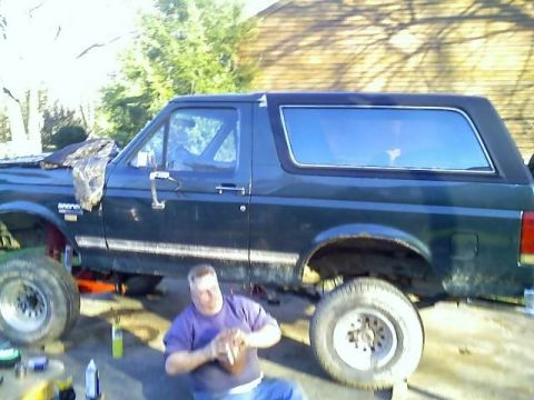 1989 Ford Bronco - A man in a purple shirt sits on the ground next to a dark green Ford Bronco, holding a football. The Bronco appears to be undergoing some m…