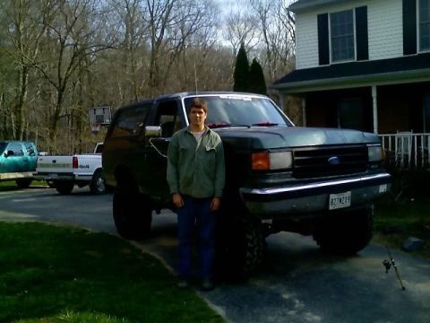 1989 Ford Bronco - A man stands in front of a lifted, dark green 1989 Ford Bronco parked in a driveway in front of a house. The Bronco has large tires and a v…