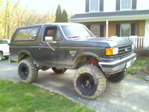 1989 Ford Bronco - A lifted, dark green 1989 Ford Bronco is parked on a paved driveway in front of a house. The vehicle has large, aggressive tires and appear…