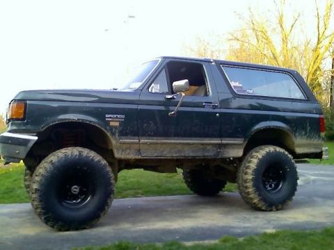 1989 Ford Bronco - A dark green, lifted Ford Bronco with large off-road tires is parked on a paved surface with grass and trees in the background. The side of…
