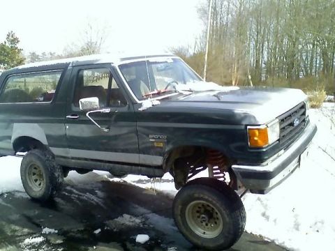 1989 Ford Bronco - A dark green Ford Bronco is parked on a snowy asphalt surface, with trees in the background. The vehicle is lifted, showing its suspension…