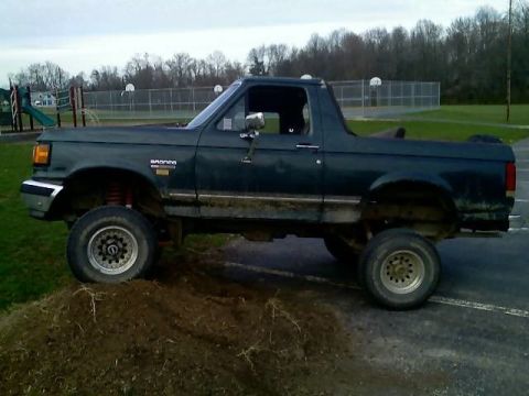 1989 Ford Bronco - A dark green, lifted Ford Bronco is parked on a dirt mound in a parking lot. The vehicle is viewed from the side with its front tires eleva…