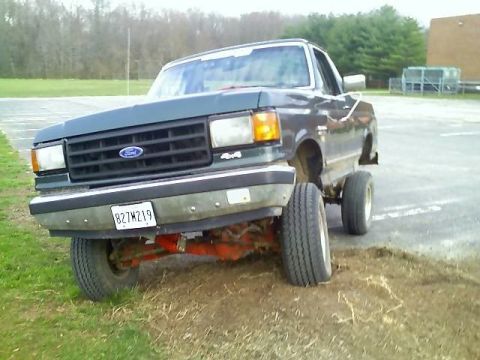 1989 Ford Bronco - A dark colored, lifted Ford pickup truck is parked at an angle on a grassy area next to a paved lot. The front of the truck is angled towar…