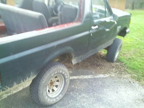 1989 Ford Bronco - The side and rear of a dark green Ford Bronco is shown parked on a gravel and asphalt surface next to a grassy area. The vehicle has a red…