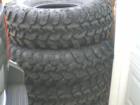 1989 Ford Bronco - A stack of three large, knobby off-road tires are leaning against a wall and a vehicle part. The tires have aggressive tread patterns suita…