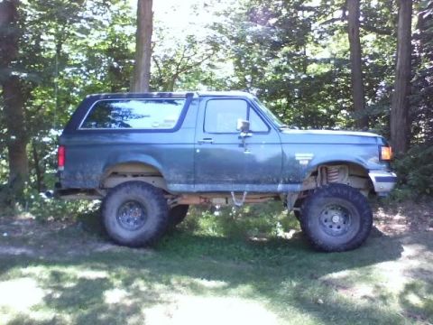 1989 Ford Bronco - A lifted, dark teal 1989 Ford Bronco with large off-road tires is parked on a grassy area surrounded by trees. The vehicle is viewed from t…