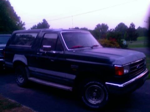 1989 Ford Bronco - The side view of a dark colored Ford Bronco is shown parked on a paved surface with trees in the background. The vehicle has a silver strip…