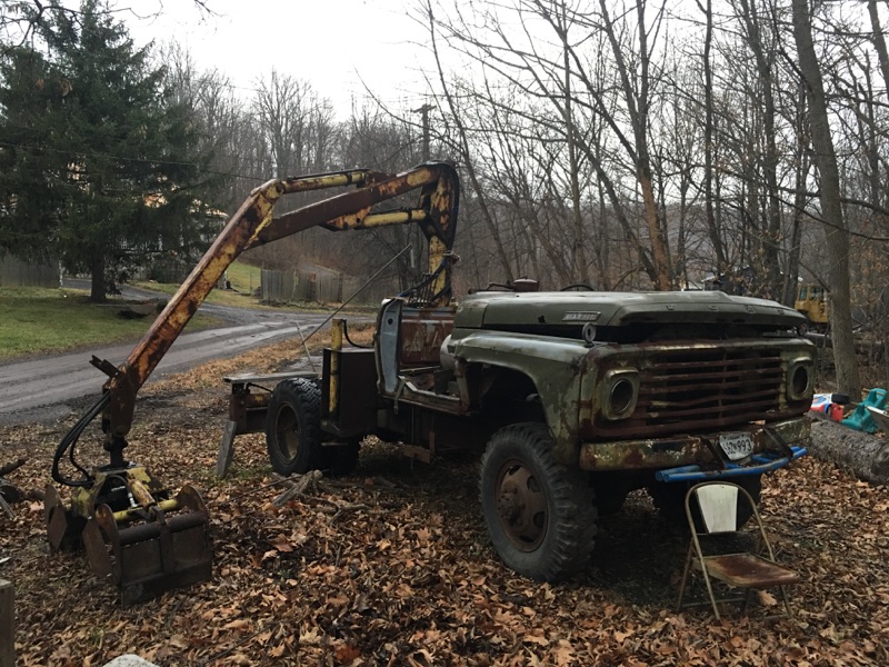 Rusty vintage Ford truck with a yellow grapple boom attachment, parked outdoors.
