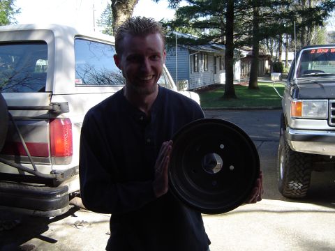 1989 Ford Bronco - Man holds a car wheel with a white Bronco and a gray truck behind him.