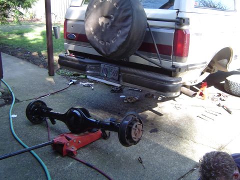 1989 Ford Bronco - The rear of a Ford Bronco is visible with a spare tire cover and bumper, along with a detached axle on a jack.