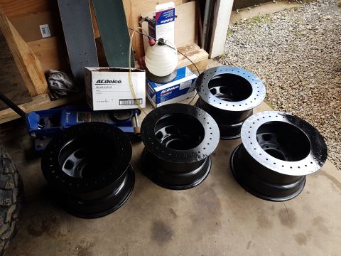 1984 Ford Bronco - Four black off-road wheels are arranged on a concrete surface, with a blue hydraulic jack and boxes of ACDelco oil in the background.