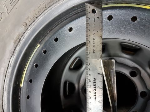 1984 Ford Bronco - A close-up view shows a metal wheel rim with a tire mounted on it, measured with a ruler. The ruler is positioned vertically, indicating a…