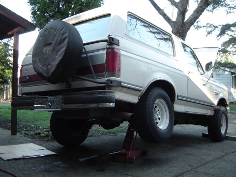 1989 Ford Bronco - A white Ford Bronco is raised on a jack, with its spare tire visible on the rear door.