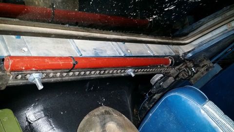 1984 Ford Bronco - A red pipe is mounted horizontally inside a vehicle, secured with zip ties and bolts. The interior features blue seats and black flooring.