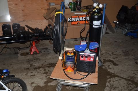 1984 Ford Bronco - A black rear axle assembly is supported by a red jack stand, with welding equipment including a helmet, welder, and gas tank nearby. The sc…