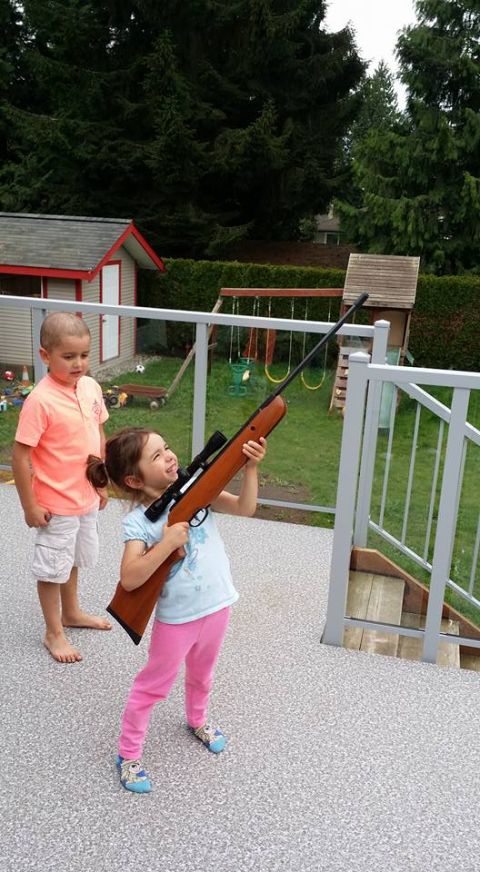 1984 Ford Bronco - A young girl holds a rifle with a scope, looking up and smiling. A young boy stands to her left, looking forward. They are on a deck with a…