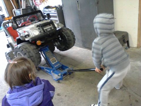 1984 Ford Bronco - A child in a hooded sweatshirt operates a blue hydraulic jack under the front tire of a silver toy SUV. Another child in a purple jacket wa…