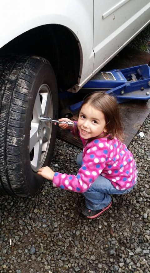 1984 Ford Bronco - A young girl in a pink polka dot jacket and jeans uses a socket wrench on the lug nuts of a vehicle's tire, which is elevated by a blue hyd…