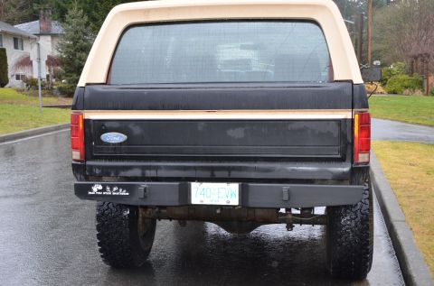 1984 Ford Bronco - The rear view of a black Ford Bronco with a tan camper shell is shown on a wet, paved road. A license plate reading "740-EVW" and a bumper…