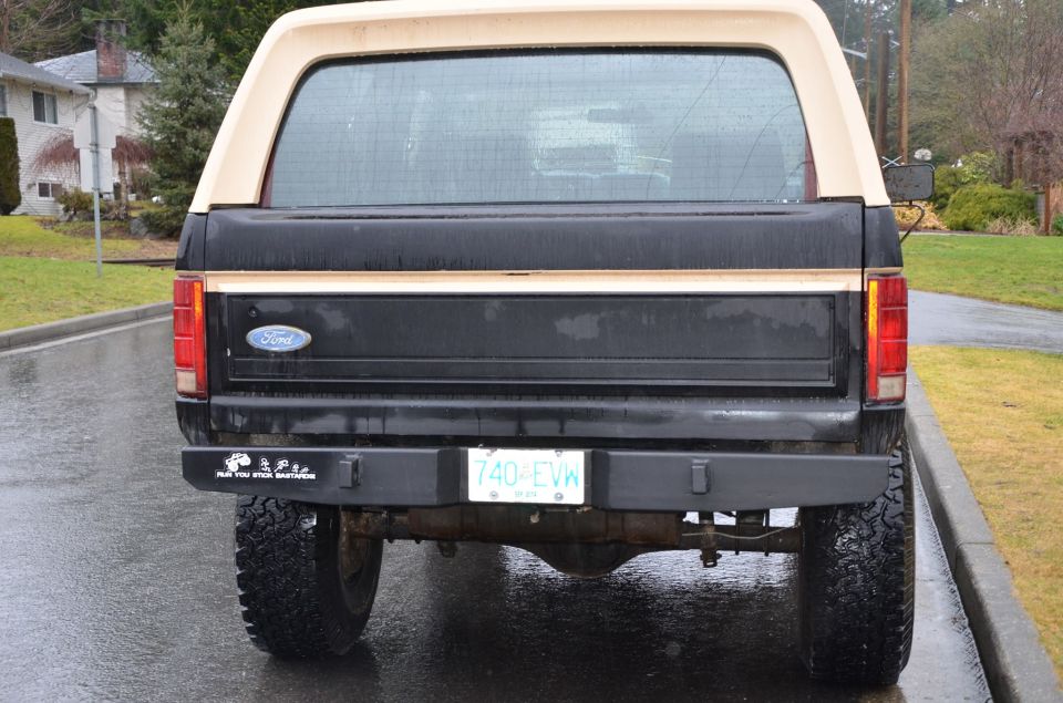 Rear view of a black 1984 Ford Bronco with a tan camper shell and a bumper sticker.