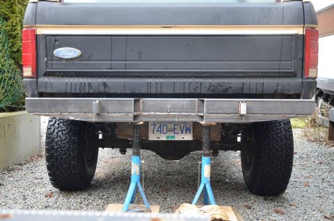 1984 Ford Bronco - The rear of a black Ford pickup truck is shown, supported by jack stands, with a custom metal bumper being installed. A license plate readi…