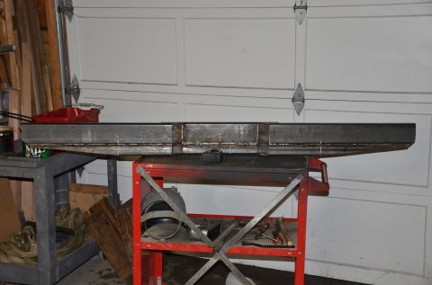1984 Ford Bronco - A long, fabricated metal beam with visible welds is resting on a red metal cart. The beam is positioned in front of a white garage door wit…