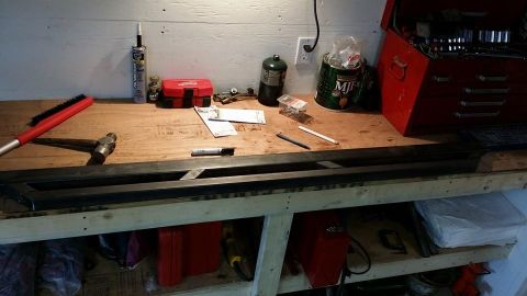 1984 Ford Bronco - A workbench is cluttered with tools and supplies, including a hammer, brush, caulk gun, propane tank, and a red toolbox. A long, dark metal…