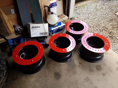 1984 Ford Bronco - Four black steel wheels with red beadlock rings are displayed on a concrete surface, with automotive fluids and boxes in the background.