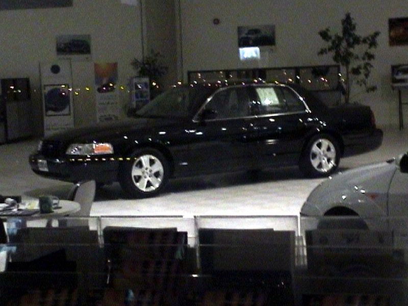 Black sedan with chrome trim and five-spoke wheels parked indoors, another car partially visible in foreground