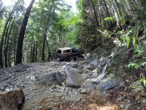 1984 Ford Bronco - A vehicle tackles a challenging, rocky off-road path through a dense forest.