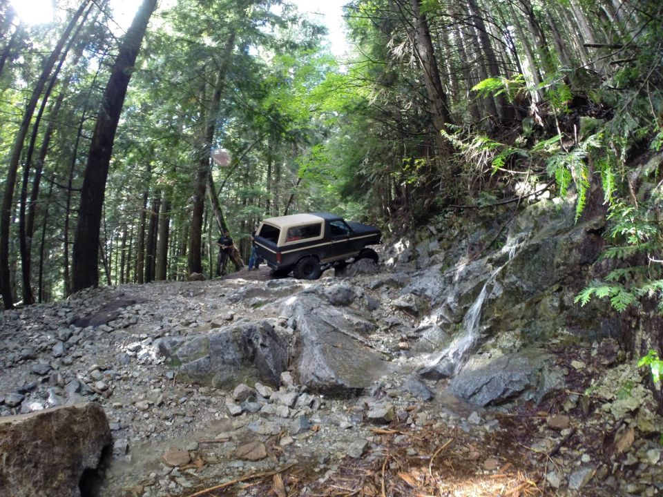 Off-road vehicle navigating a steep, rocky trail with trees on either side. Water is flowing down the rocks