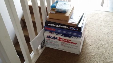 1984 Ford Bronco - Boxes for B&M automotive parts, including a torque converter and transmission cooler, are stacked on a carpeted floor near stairs.