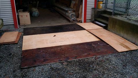 1984 Ford Bronco - Plywood sheets are arranged to create a makeshift ramp over gravel and a dark surface in front of a garage.