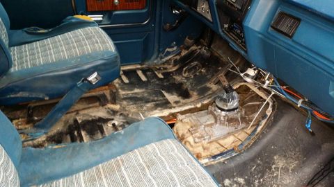 1984 Ford Bronco - The interior of a vehicle shows a dirty floor and exposed transmission components, with blue plaid seats.