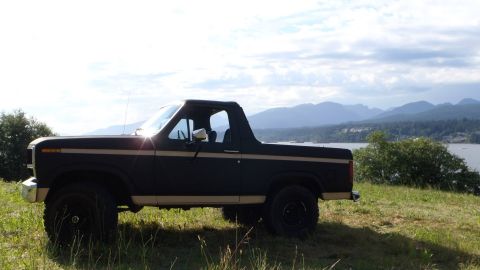 1984 Ford Bronco - Ford Bronco parked on a hill overlooking a body of water and mountains under a cloudy sky.