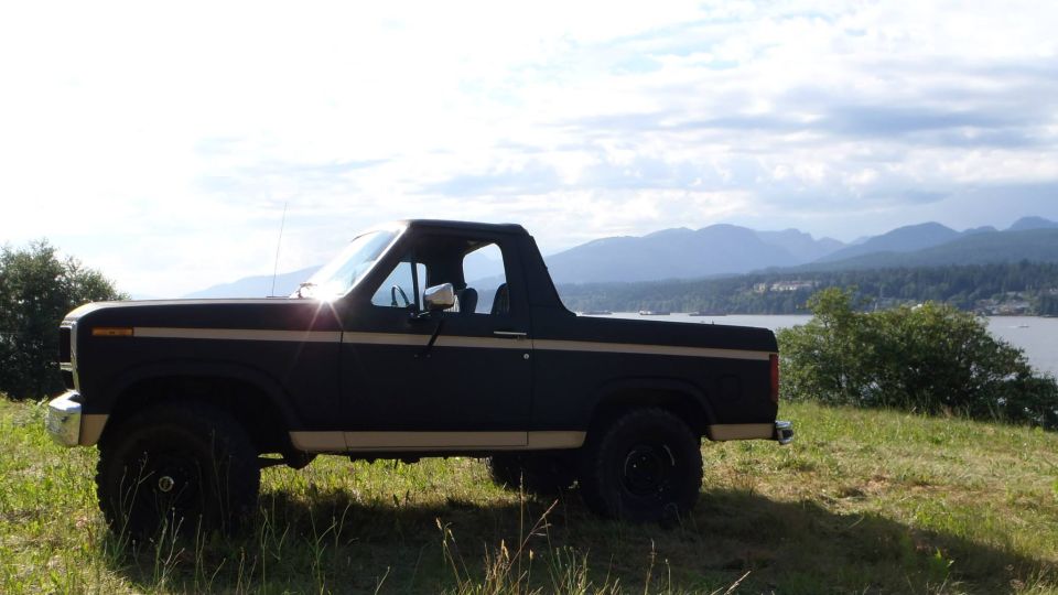 Side view of a dark 1980s Ford Bronco with tan pinstripes, parked on a hill with a bay and mountains in the background