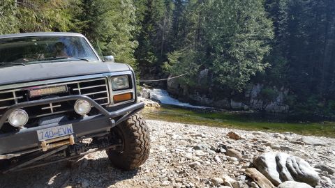 1984 Ford Bronco - Off-road SUV parked on a rocky shore near a waterfall in a wooded area.