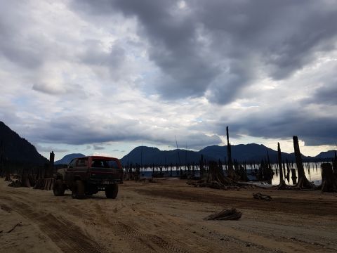 1984 Ford Bronco - Ford Bronco parked on a dirt road beside a lake with dead trees under a cloudy sky.