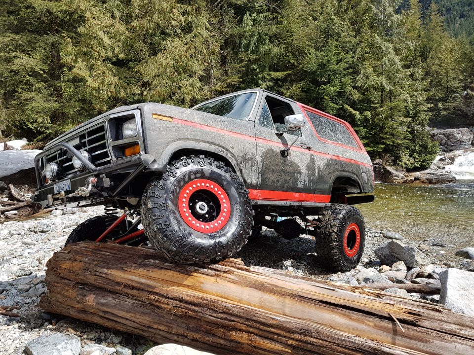 Modified Ford Bronco with large tires and red rims on a log near a river